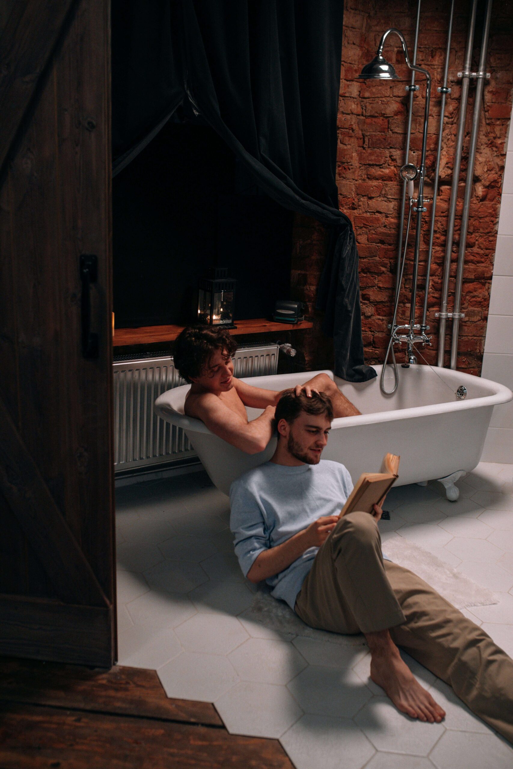 male gay couple in the bathroom, one in the tub and the other closeby reading a book